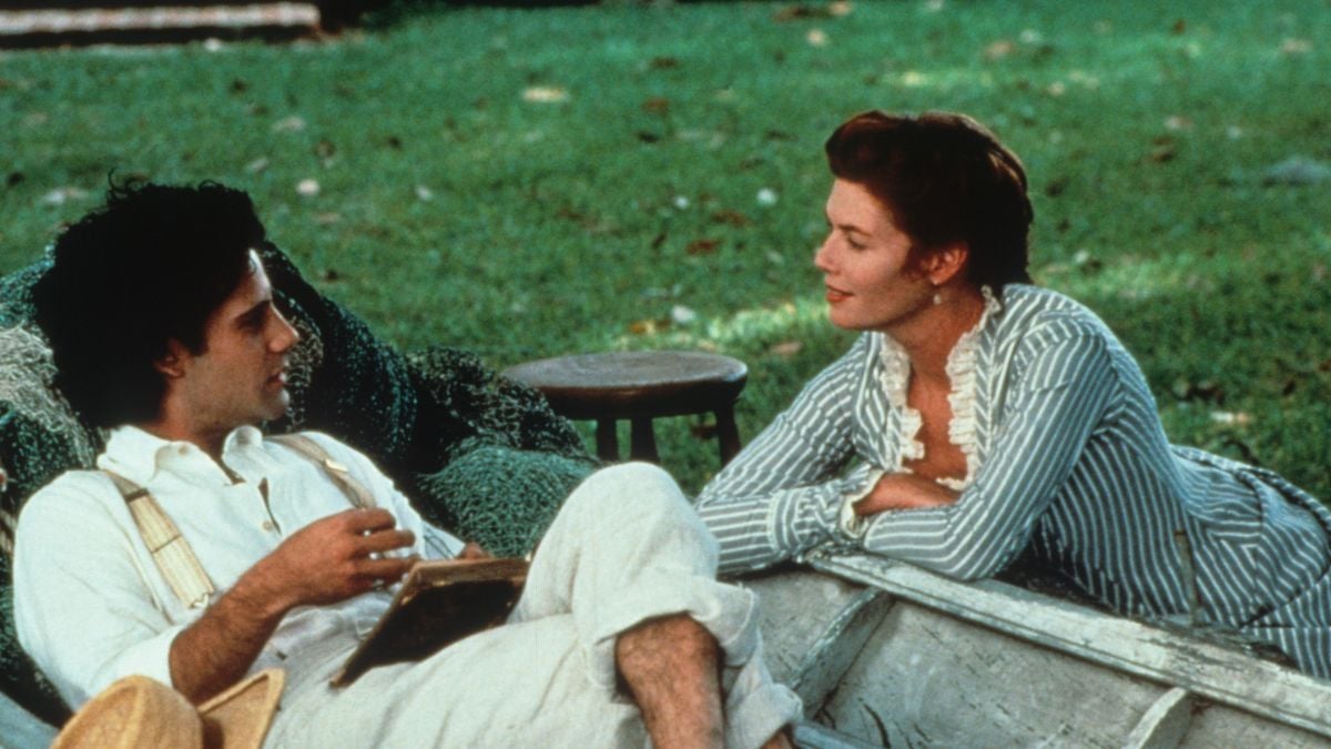 A man and woman relax outdoors on a blanket, facing each other and talking. The man holds a book, while the woman leans in, smiling gently. Lush green grass and sunlight surround them.