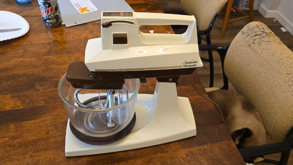 A vintage white and brown Sunbeam Mixmaster stand mixer with a glass bowl on a wooden table.