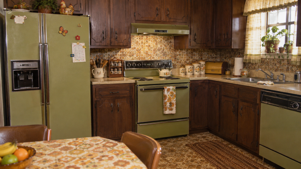 Vintage kitchen with avocado green appliances, dark wood cabinets, floral wallpaper, a round table with a patterned tablecloth, and potted plants on the window sill. Sunlight streams through a lacy curtain.