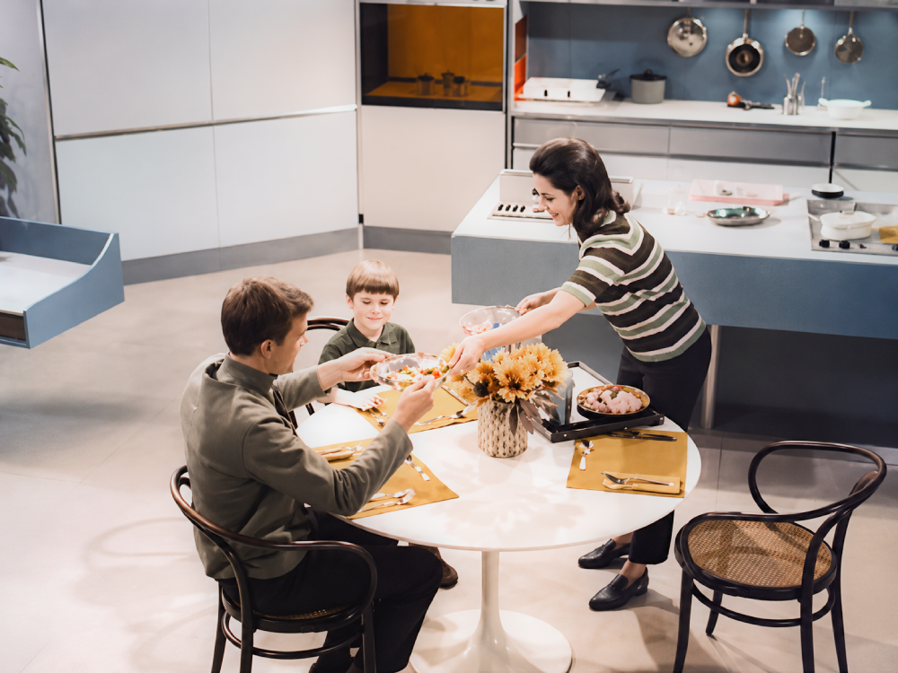 A woman in a striped shirt serves a plate of food to a man and a young boy sitting at a round dining table in a kitchen.