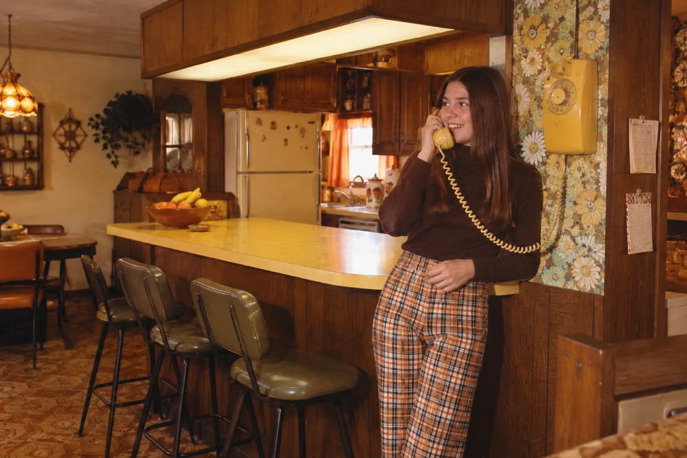 A woman in plaid pants talks on a yellow rotary phone in a vintage kitchen with wood cabinets, floral wallpaper, and retro decor.