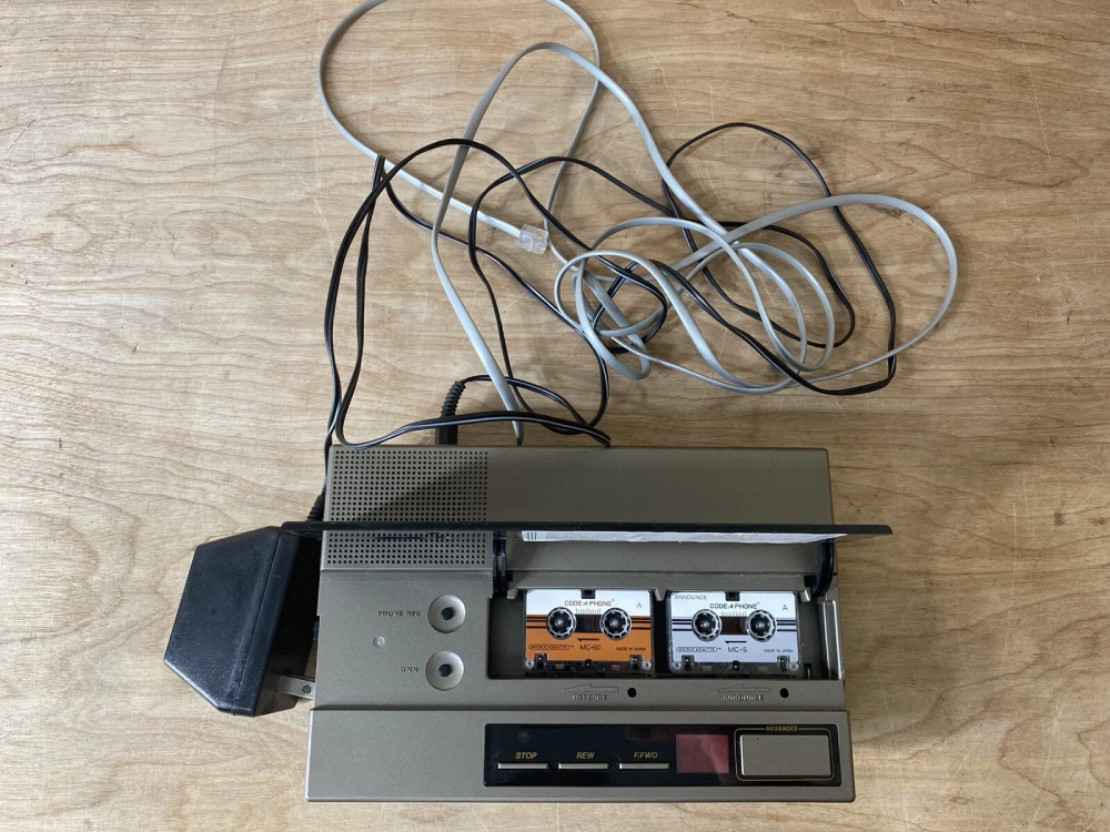 A vintage Code-A-Phone answering machine with two microcassette tapes inserted, connected to tangled power and telephone cords.