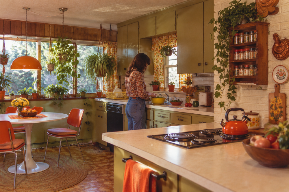 A woman stands in a retro kitchen with green cabinets, orange decor, and lots of plants. Sunlight streams in from large windows above the sink. A table with flowers and orange chairs sits nearby.