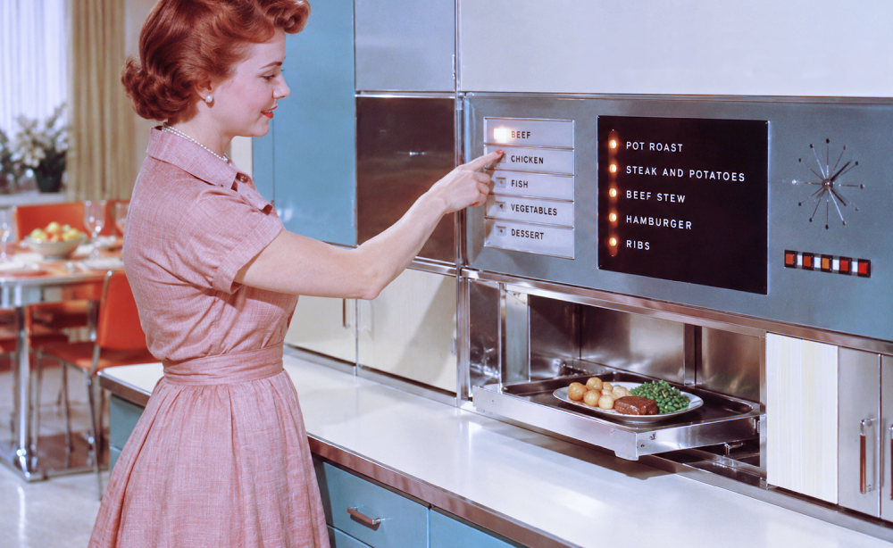 A woman in a pink dress presses a button on a futuristic kitchen appliance labeled with food categories and meal options, with a plate of food sitting in the open warming drawer.