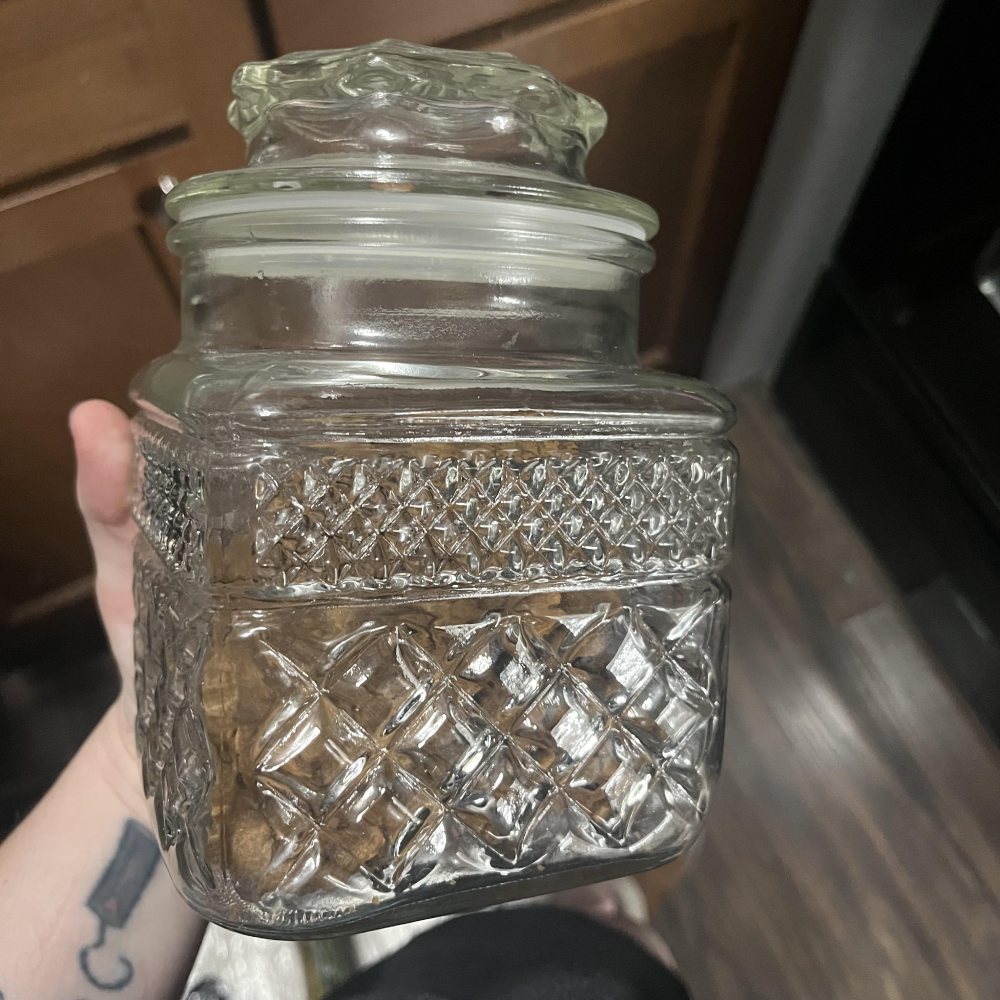 A hand holds a square glass storage jar with a diamond-patterned texture and a glass lid.