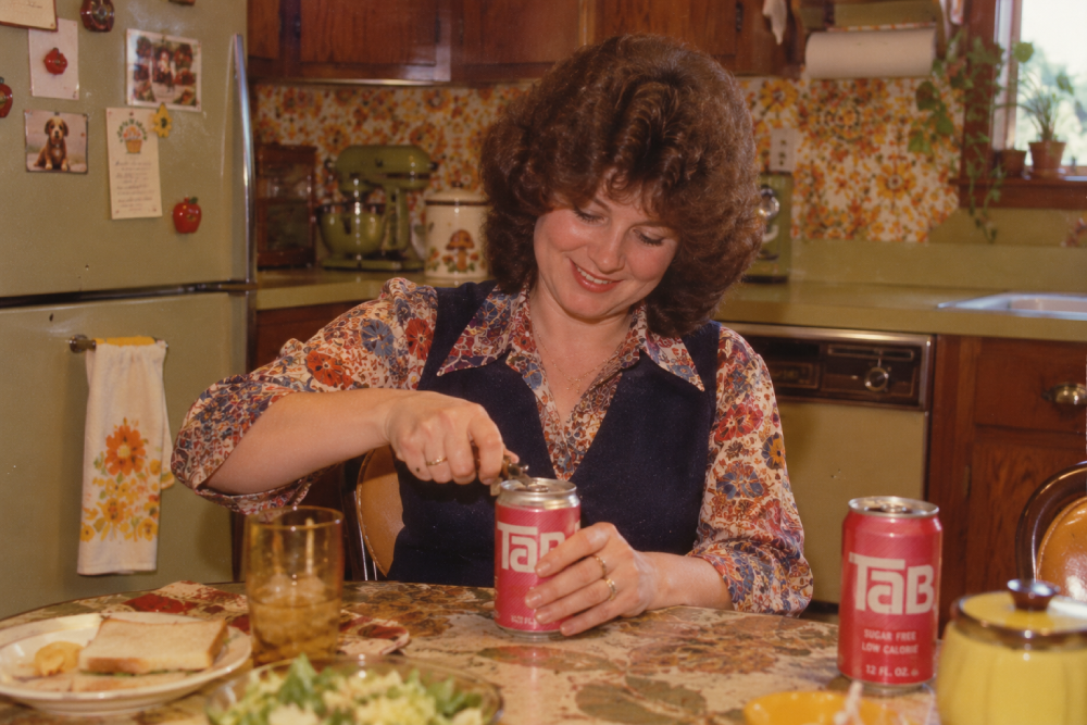 A woman in a vintage kitchen smiles while opening a can of Tab soda. Another Tab can and plates of food are on the table. The kitchen has floral wallpaper, wooden cabinets, and an old refrigerator.