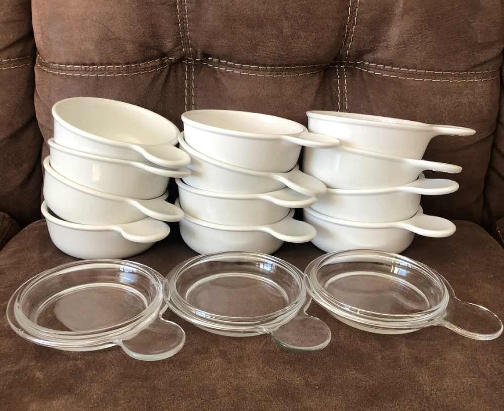 Nine white ceramic bowls with handles stacked in three piles, with three clear glass lids with handles in front.