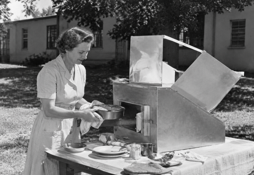 A woman in a dress places a round baking pan into a box-shaped solar oven on an outdoor table.