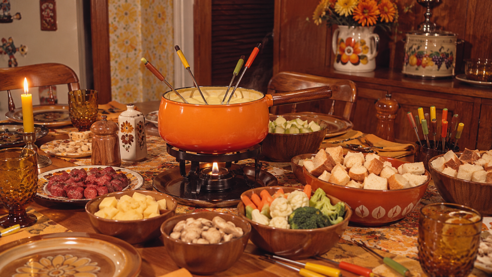 A retro fondue dinner setup with an orange pot of melted cheese, skewers, and bowls of bread, meat, vegetables, and fruit on a vintage table with candles and 1970s-style decor.