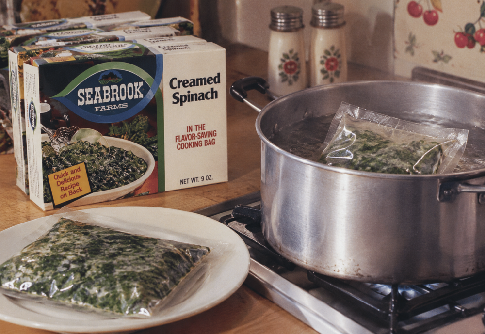 A box of Seabrook Farms creamed spinach next to a plate holding a frozen pouch and a pot on the stove containing another pouch of spinach.