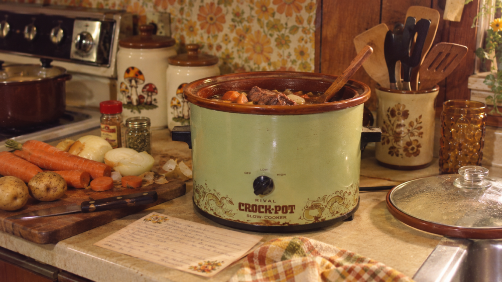 A vintage slow cooker with stew sits on a kitchen counter surrounded by fresh vegetables, spices, a recipe card, and utensils, creating a cozy, retro cooking scene.
