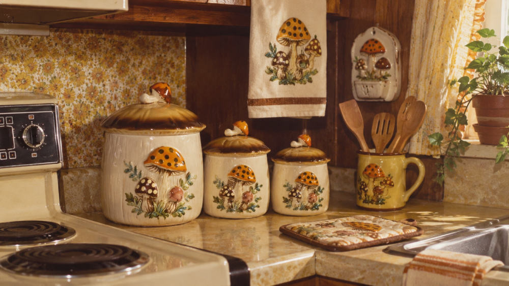 Vintage kitchen with mushroom-themed canisters, towel, oven mitt, and utensil holder on a yellow counter. Floral wallpaper, stove, sink, and potted plant are also visible. Warm, retro atmosphere.