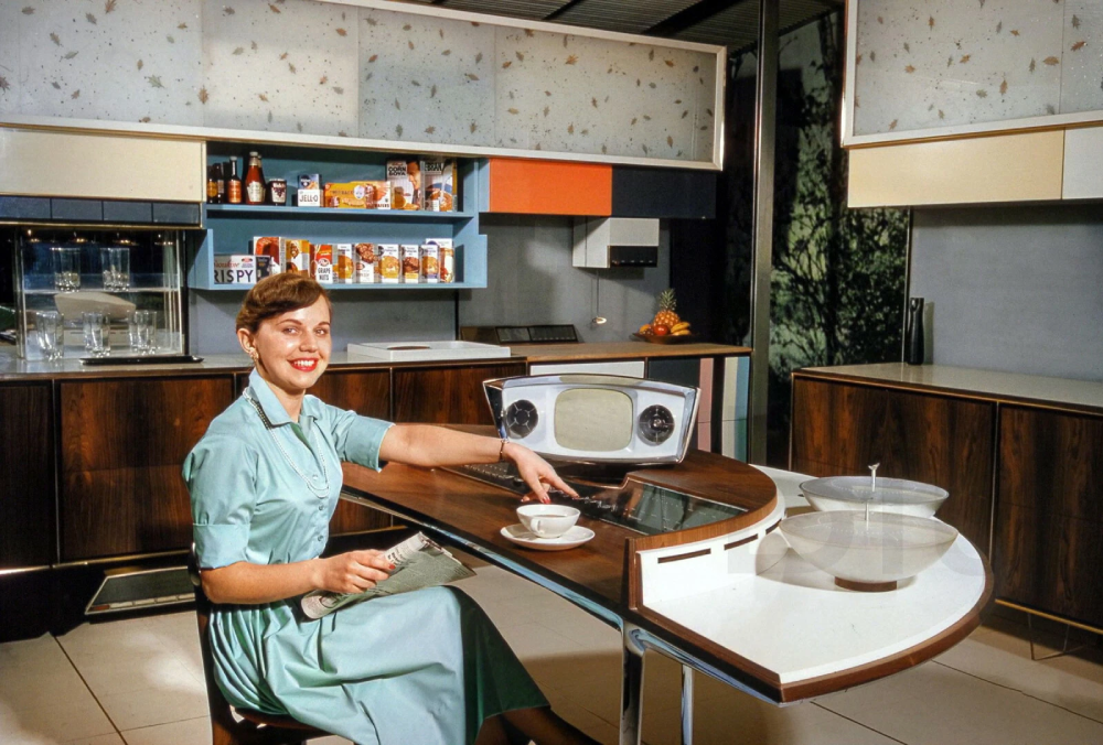 A woman in a light blue dress sits at a futuristic kitchen island with a built-in television, holding a magazine and a teacup.