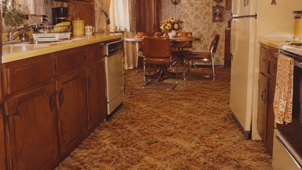 1970s-style kitchen with brown cabinets, yellow countertops, floral wallpaper, patterned carpet, and a dining area with brown chairs and a round table decorated with flowers.