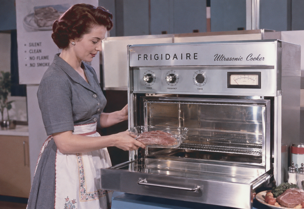 A woman in an apron places a glass dish of meat into a Frigidaire Ultrasonic Cooker.