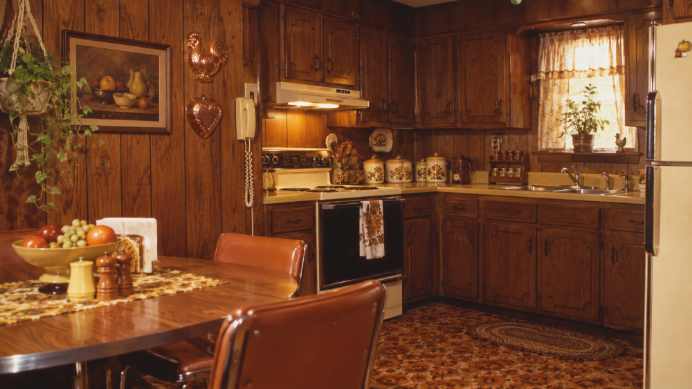 A vintage kitchen with dark wood cabinets, yellow appliances, a round table with brown chairs, plants, and decorative jars. The decor includes a fruit painting and patterned carpet.