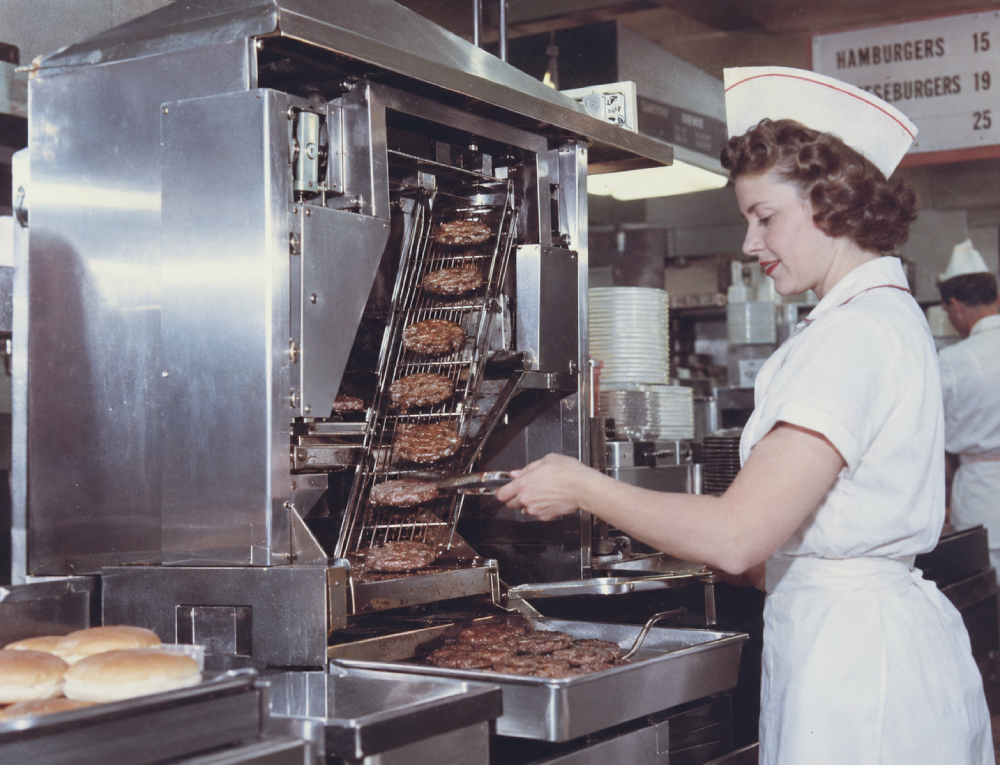A woman in a white uniform and cap uses tongs to remove cooked hamburger patties from a vertical conveyor-belt grill.