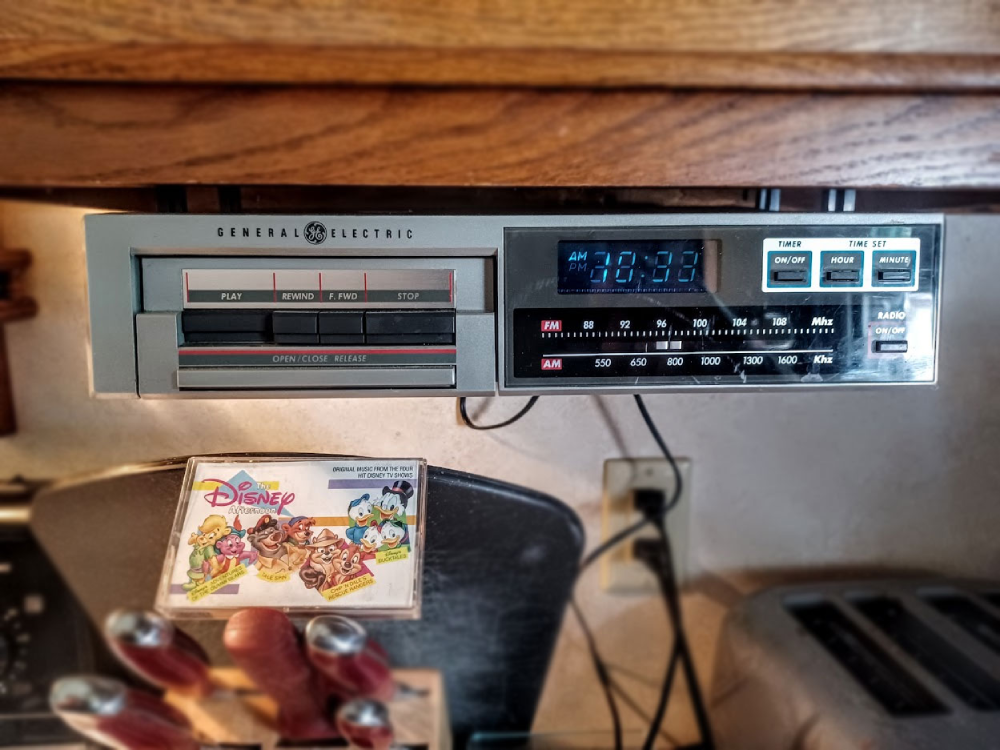 A General Electric under-cabinet radio and cassette player mounted below a kitchen cupboard, with a Disney Afternoon cassette tape sitting in the foreground.