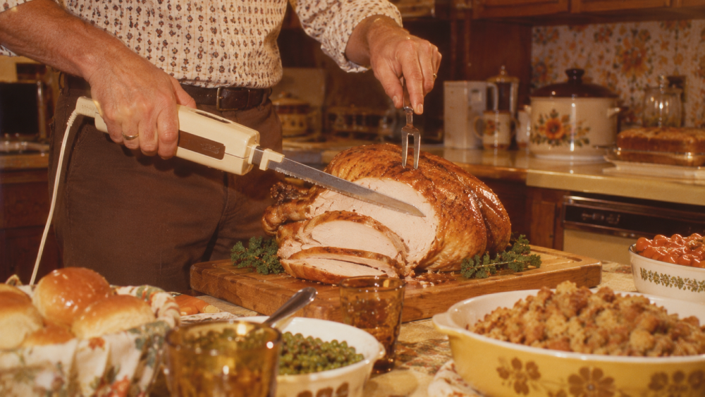 A person slices a roasted turkey with an electric knife on a wooden board, surrounded by bowls of stuffing, green peas, dinner rolls, and other dishes in a cozy kitchen setting.
