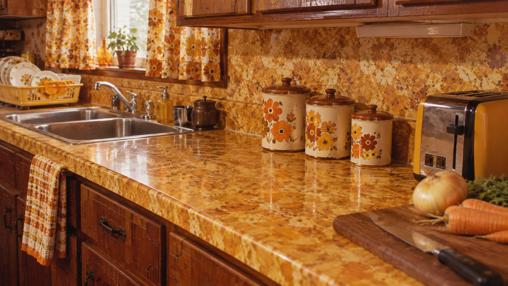 A retro kitchen with brown cabinets, a double sink, yellow floral canisters, a toaster, and vegetables on a cutting board. The counters and backsplash are yellow-gold with a marbled pattern.