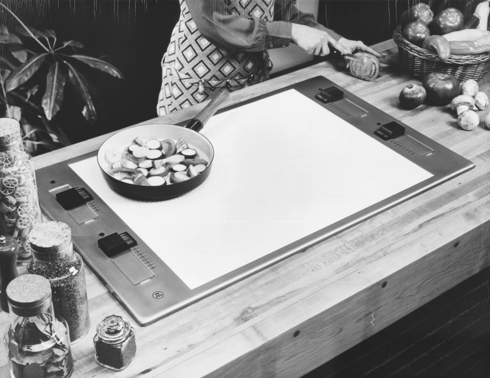 A person slices vegetables on a kitchen counter next to a built-in electric cooktop with a frying pan.