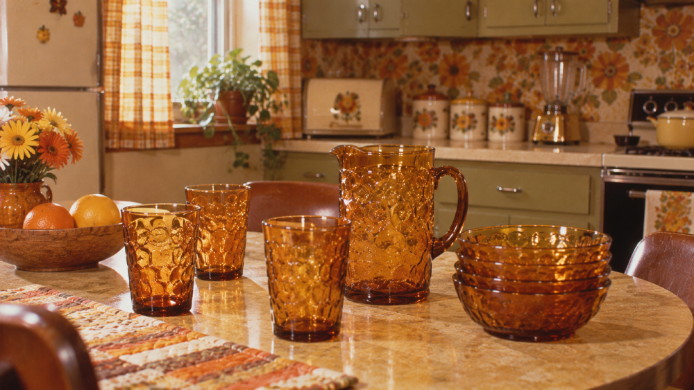 Amber glass pitcher, cups, and bowls on a kitchen table with a bowl of fruit and flowers; retro kitchen with green cabinets, patterned wallpaper, and sunlight through a window in the background.