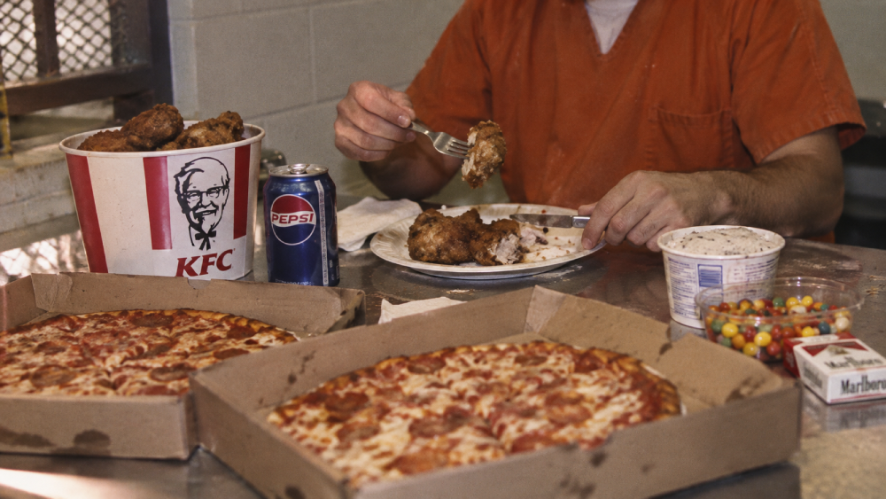 A person in an orange prison uniform eats fried chicken at a table covered with two pepperoni pizzas, a bucket of KFC, a Pepsi can, a small tub of side dish, a container of candies, and a pack of cigarettes.