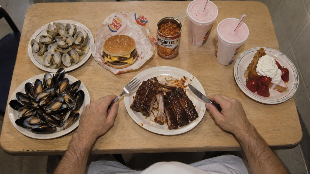 A first-person view of a table filled with various foods, including bowls of clams and mussels, a double cheeseburger, a can of SpaghettiOs, two milkshakes, a slice of strawberry pie, and a plate of barbecue ribs being eaten with a knife and fork.