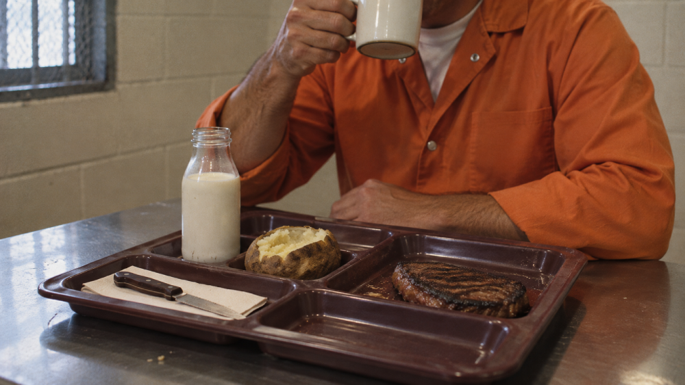 A person in an orange jumpsuit sits at a table drinking from a mug, with a prison-style cafeteria tray holding a steak, a baked potato, and a small bottle of milk.