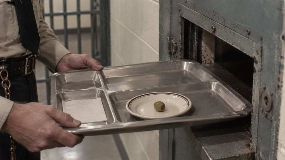 A prison guard slides a metal tray containing a single olive on a small plate and a napkin through a slot in a cell door.