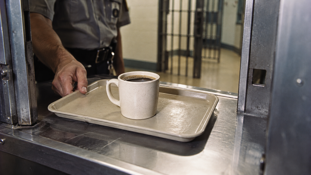 A uniformed officer slides a tray with a mug of coffee through a slot in a jail cell door.