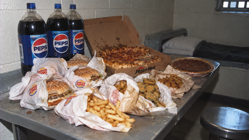A metal table in a jail cell covered with Burger King burgers and fries, a Pizza Hut pizza, three large bottles of Pepsi, and a pie.