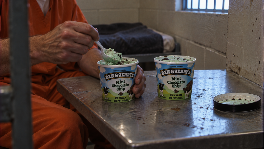 A person in an orange prison uniform eats Ben & Jerry’s Mint Chocolate Chip ice cream from a pint container at a metal table.