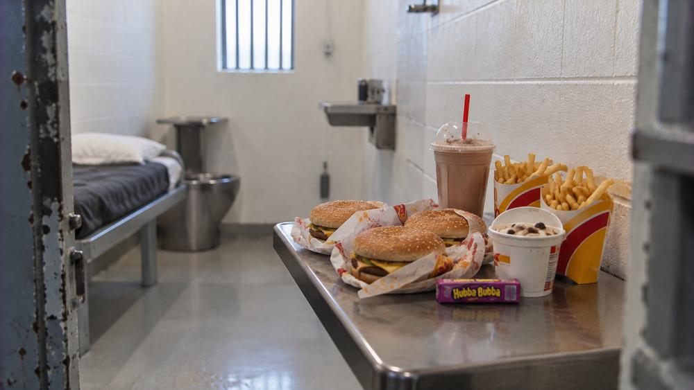 A metal table inside a prison cell holds a fast-food meal of three burgers, two orders of french fries, a chocolate milkshake, a cup of ice cream, and a pack of Hubba Bubba gum.