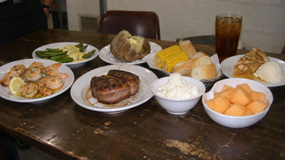 A table set with a meal including shrimp, asparagus, a baked potato, steak, corn on the cob, bread, cottage cheese, melon, apple pie with ice cream, and a glass of iced tea.