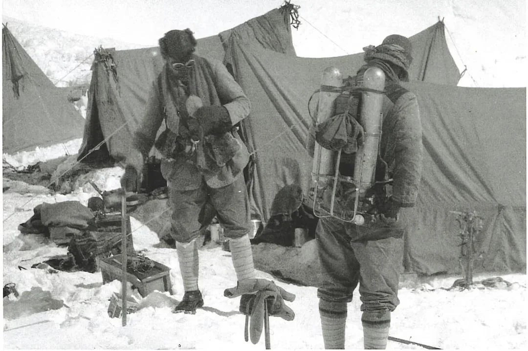 Two mountaineers in cold-weather gear stand outside tents in the snow, with one climber wearing an oxygen apparatus on his back.