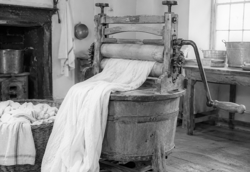 A vintage wooden clothes wringer mounted on a laundry tub with a cloth being fed through the rollers.