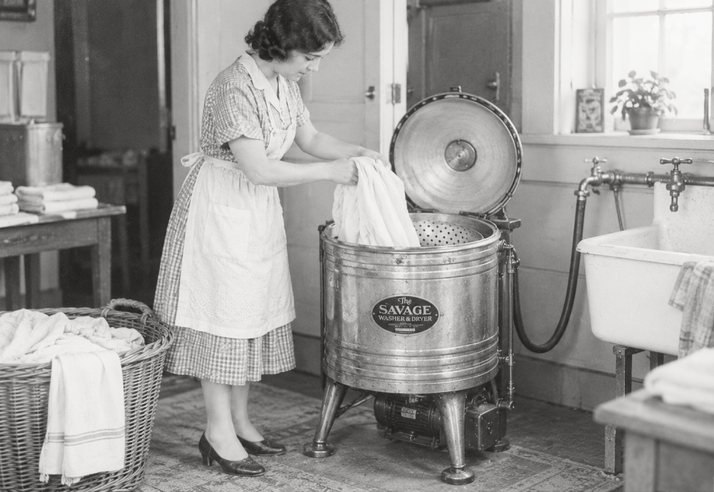 A woman in a dress and apron loads clothing into a metal Savage brand washing machine.