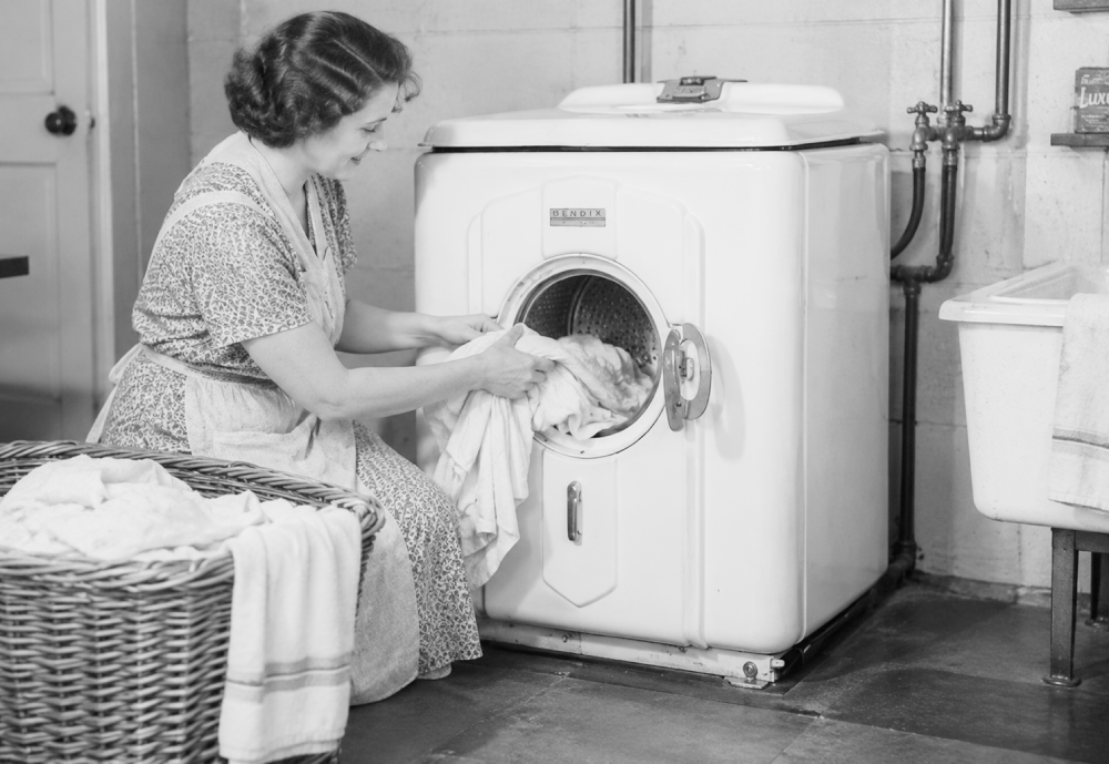 A woman in an apron loads laundry into a front-loading Bendix washing machine next to a wicker laundry basket.