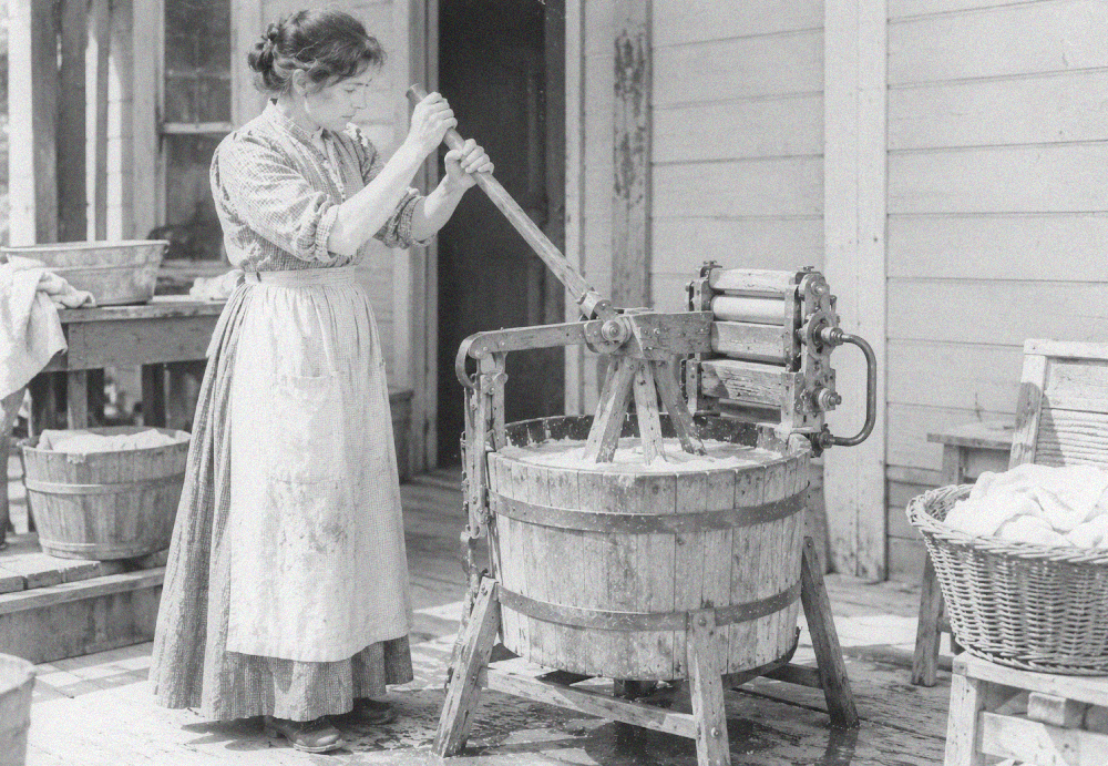 A woman in an apron uses a hand-cranked wooden washing machine on a porch.