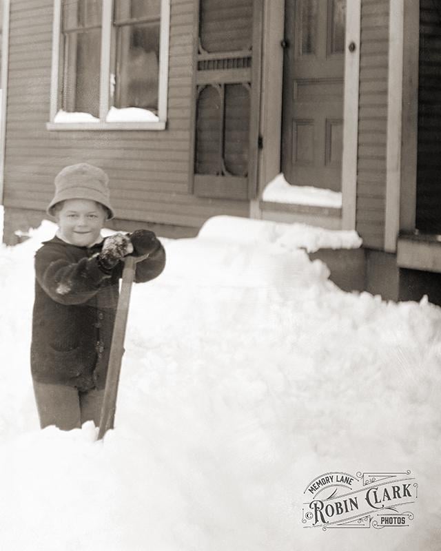 A child wearing a hat and coat shovels deep snow in front of a house.