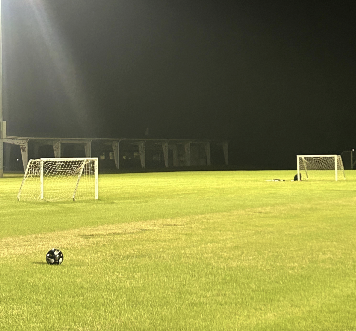 A soccer ball sits on a field at night with two goals visible under stadium lights.