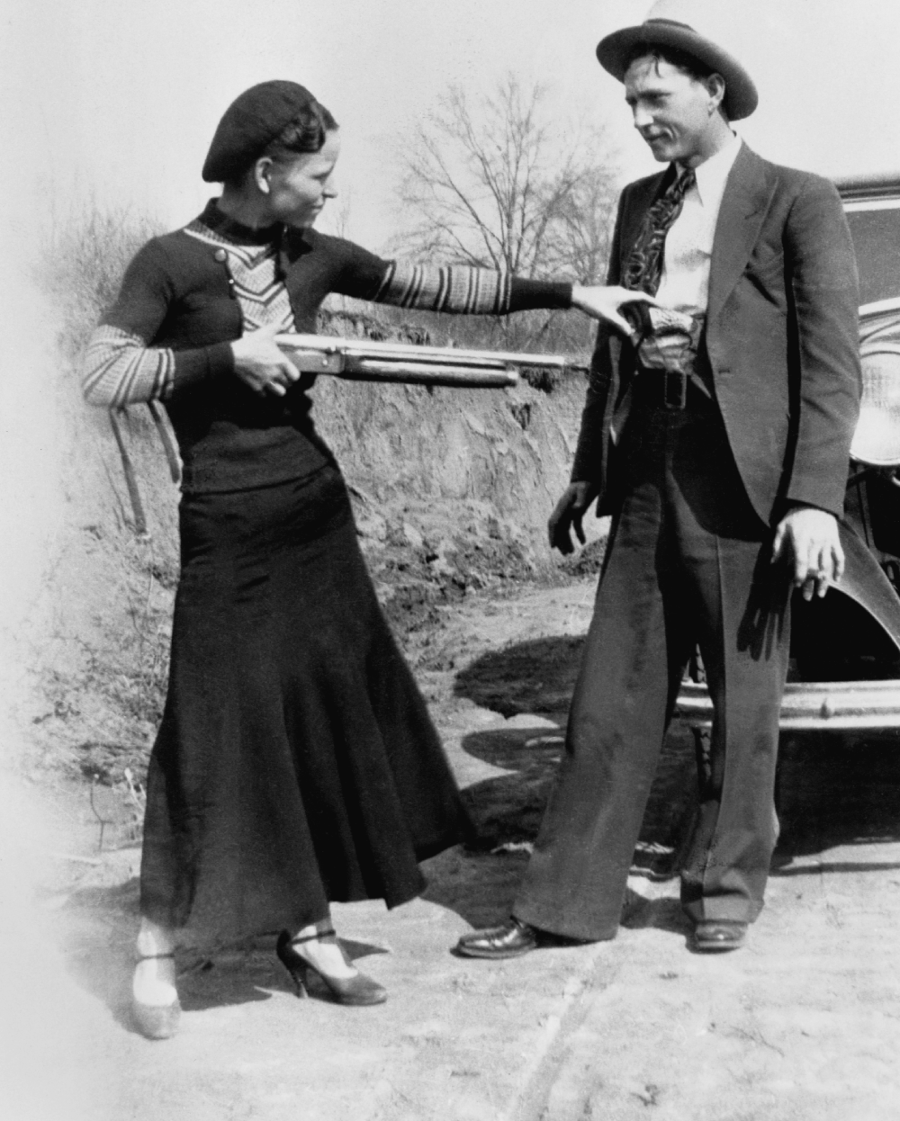A woman in a skirt and beret points a shotgun at the chest of a man in a suit and fedora standing by a car.