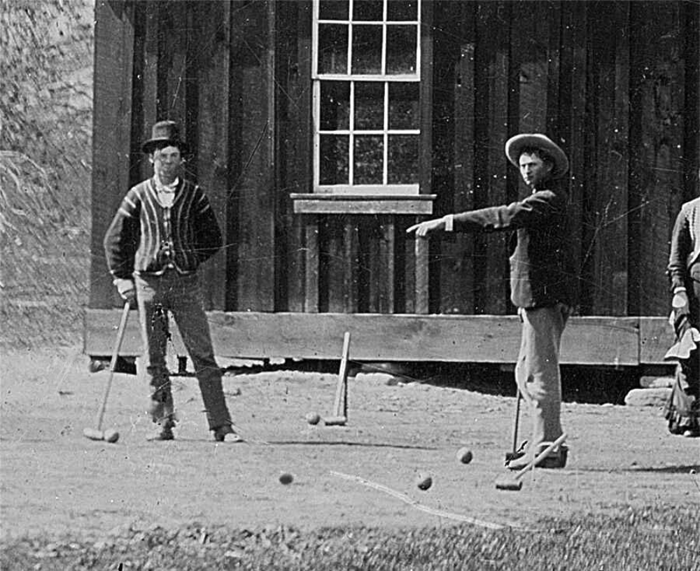 Two men in hats and period clothing playing croquet in front of a wooden building.