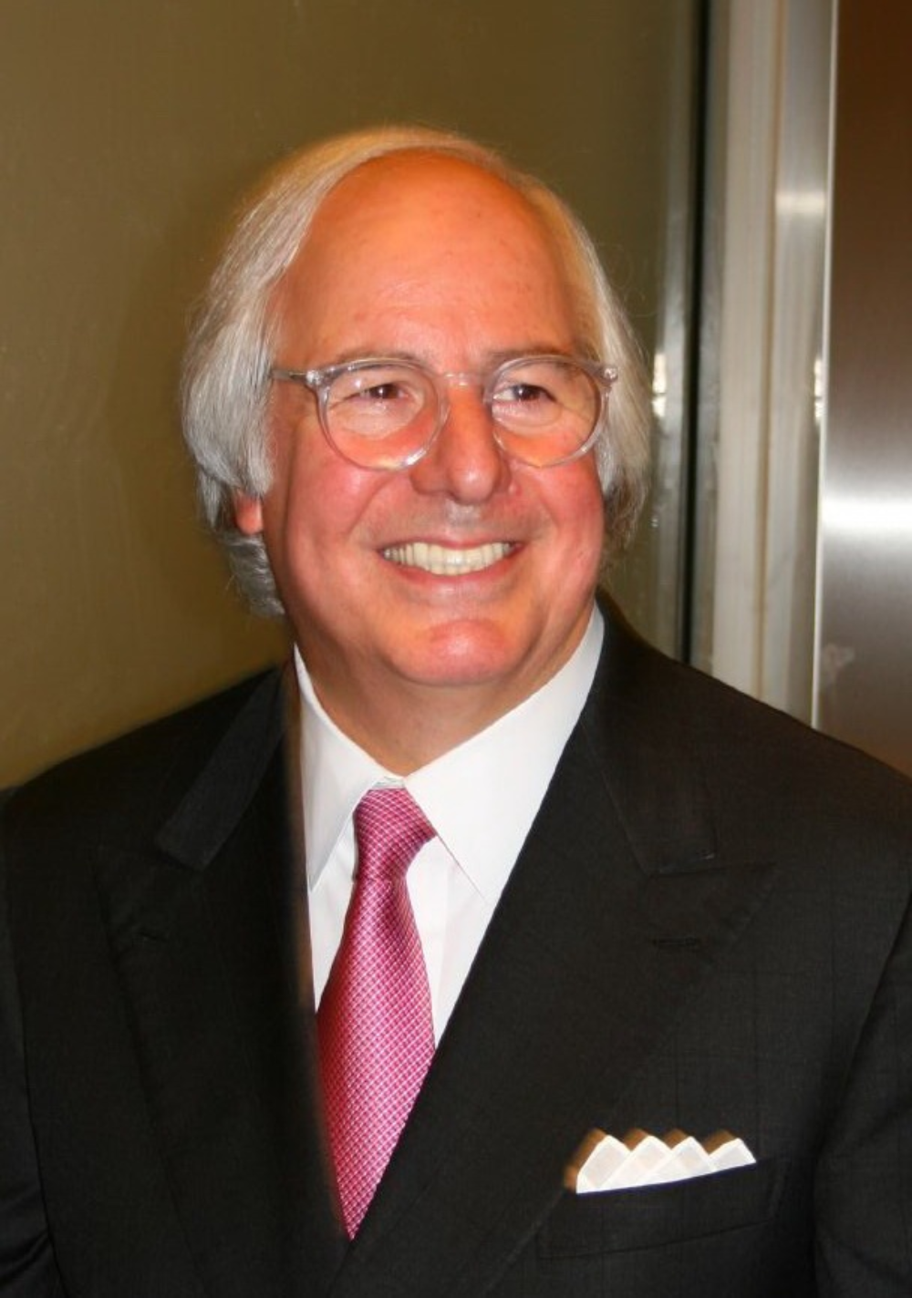 Middle-aged man with white hair wearing clear-rimmed glasses, a dark suit, white shirt, pink tie, and a white pocket square.