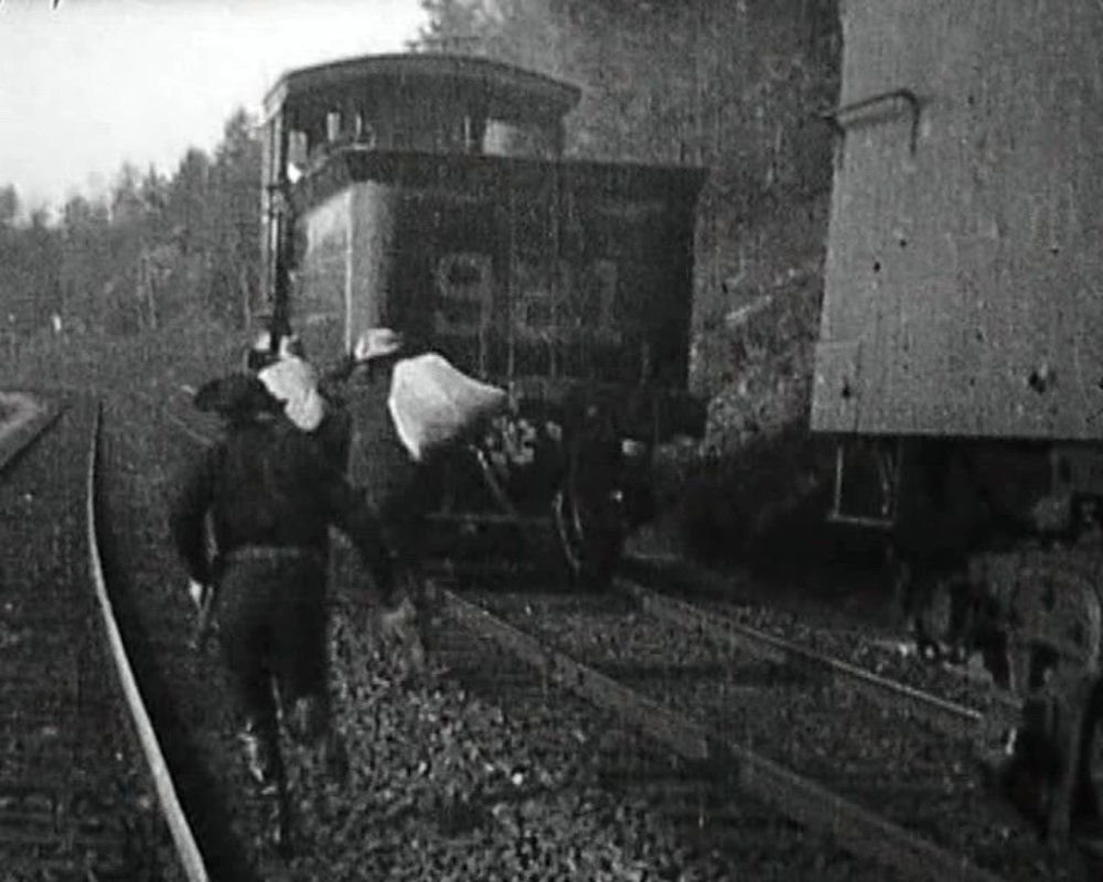 Two people carrying bags run alongside a moving train on a railway track.