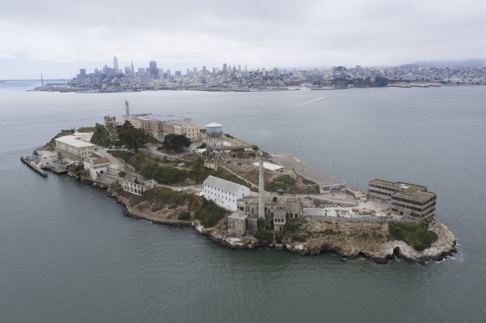 Aerial view of Alcatraz Island, featuring its historic prison buildings, with the San Francisco skyline visible across the bay in the background.
