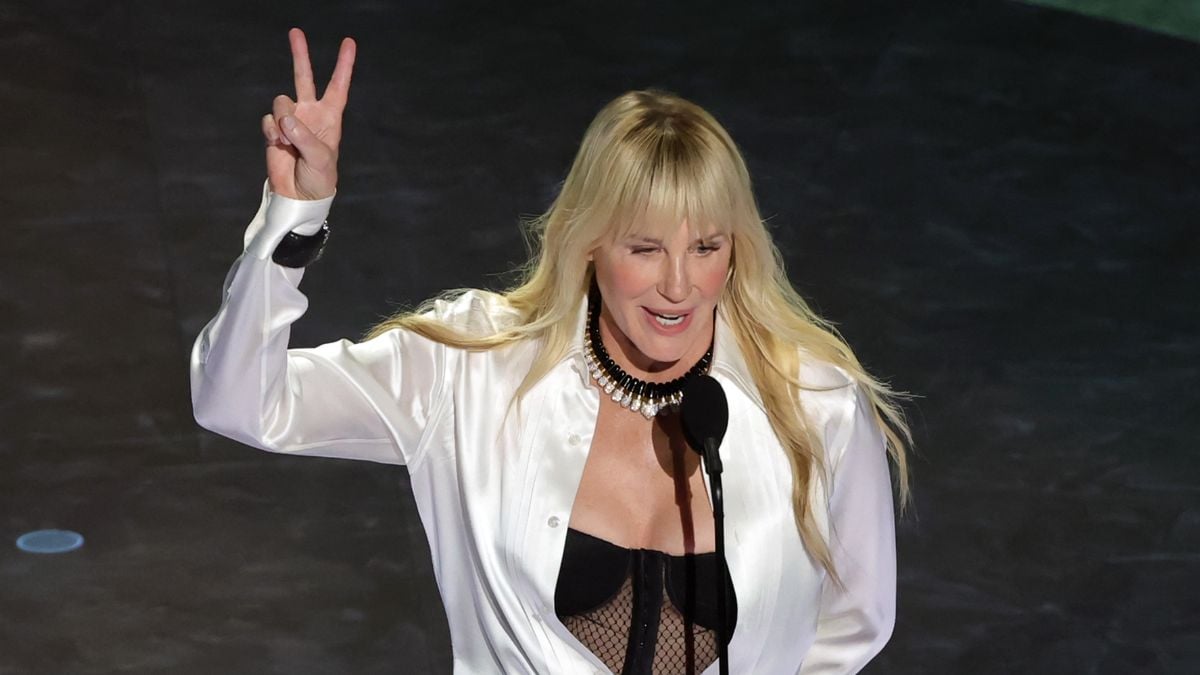 A woman with long blonde hair wearing a white shirt and black top speaks at a microphone, raising her hand and making a peace sign gesture.