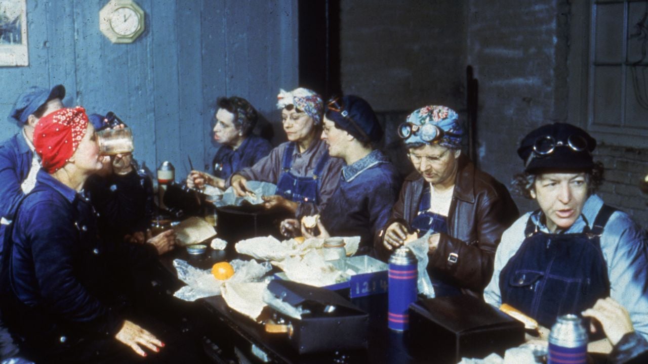 A group of women in work uniforms and bandanas sit at a table, eating lunch together. Lunchboxes and thermoses are visible in front of them. The setting appears to be a break room or industrial workplace.