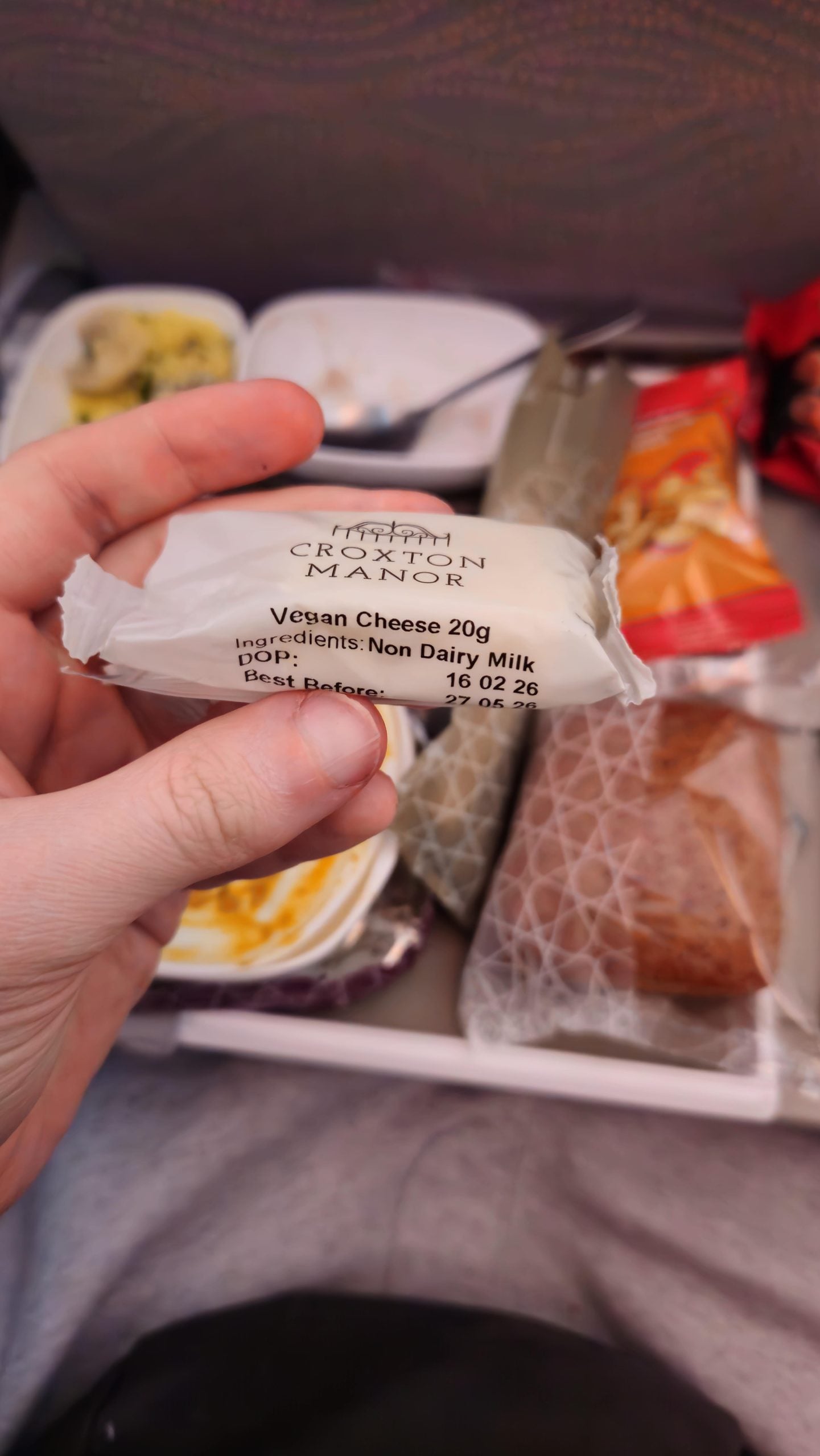 A hand holds a small, individually wrapped portion of Croxton Manor vegan cheese over an airplane meal tray.